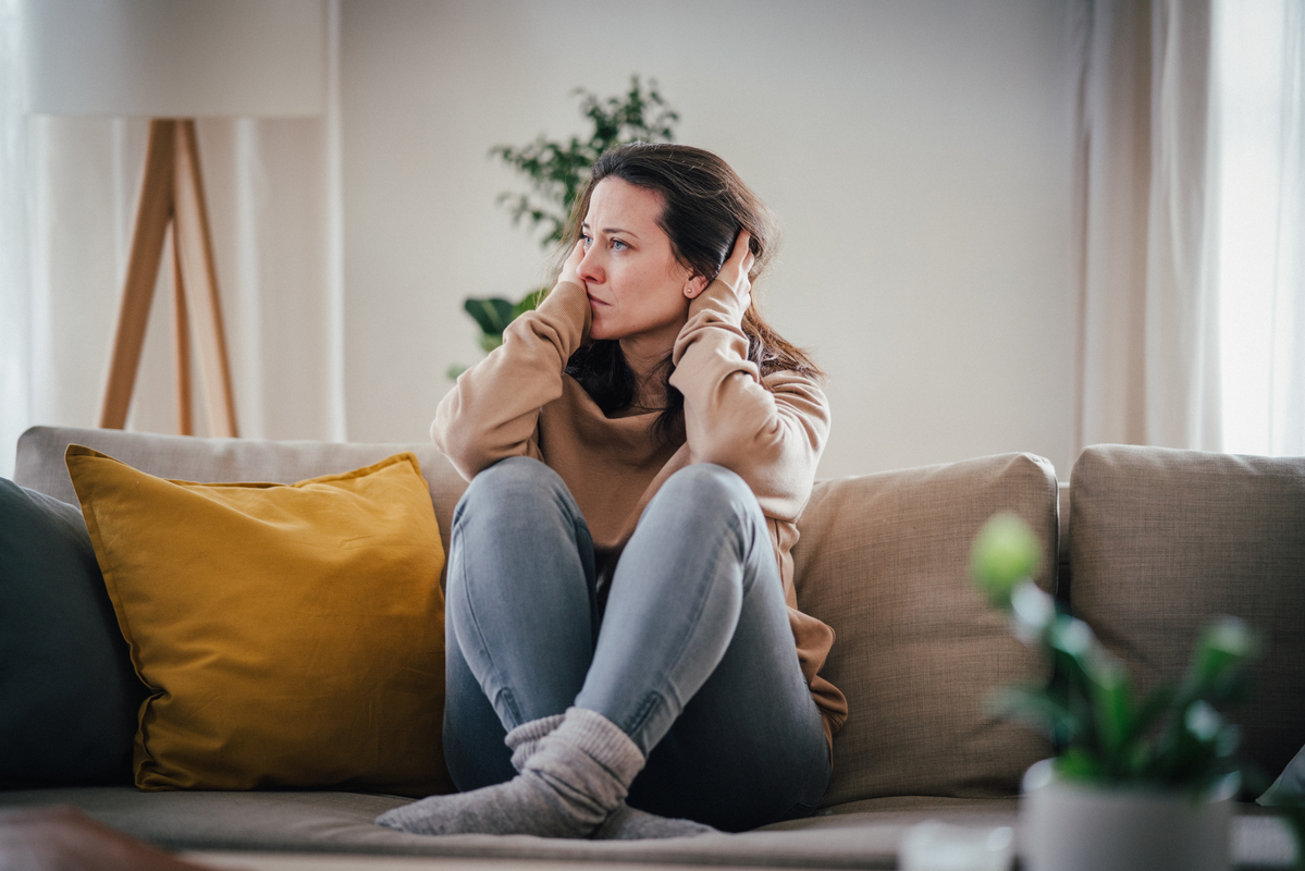 mladá žena sedí na gauči se zkříženýma nohama, drží se rukama za hlavu a vypadá velmi ustaraně a unaveně