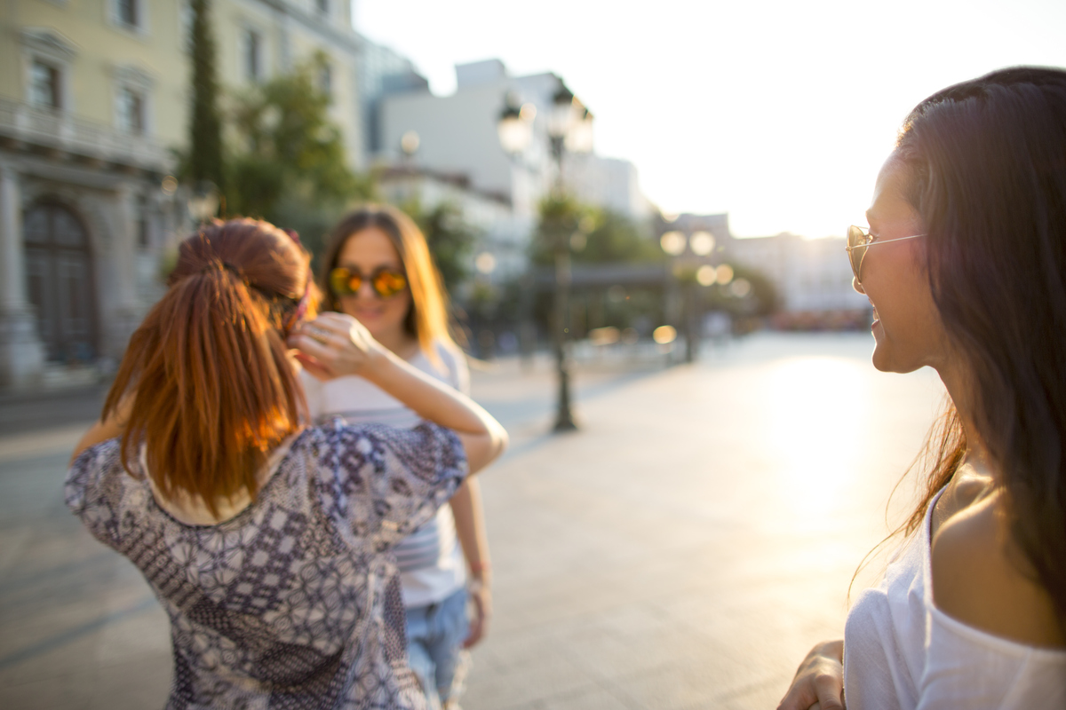 Tři ženy se slunečními brýlemi na ulici v dobré náladě si povídají, jedna z nich je zády a vidíme tak pouze její zdravé vlasy