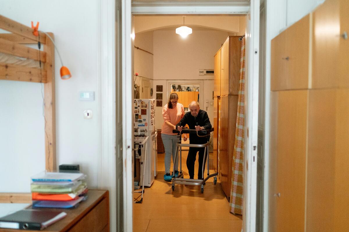 pečovatelka pomáhá seniorovi s chůzí v jeho bytě