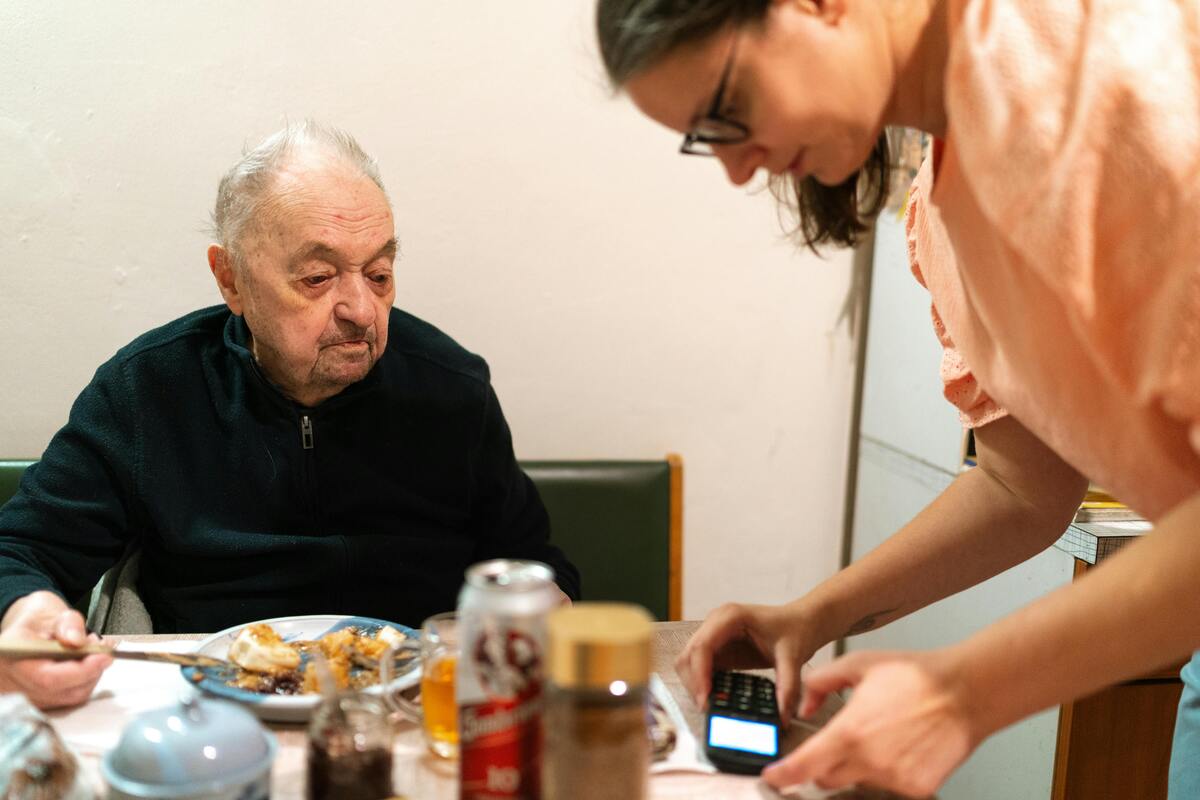 pečovatelka pomáhá seniorovi s nastavením telefonu, zatímco on večeří